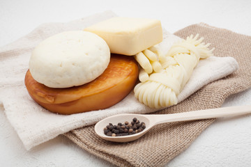 Types of cheese, including smoked, hard, sour-milk and pigtail, are decorated with black pepper in polka dots, located on a decorative tablecloth, on a white background. The concept of cheese.