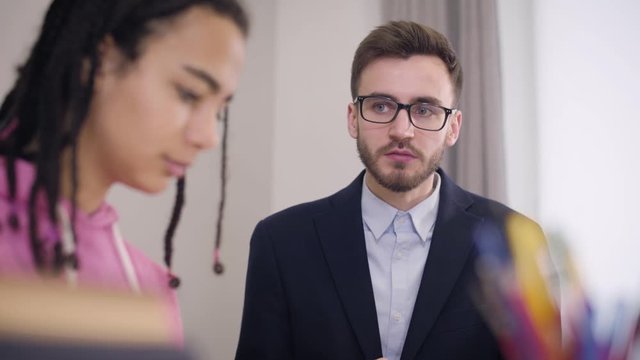 Close-up of strict male Caucasian teacher scolding young African American university student. Blurred sad girl at the foreground listening to tutor carefully. Education, problems, diligence.