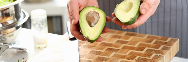 Male Hands Holding Green Avocado Fruit Halves. Chef in Apron Showing Cut Tropical Ingredient with Brown Seed. Man Cooking Food on Wooden Board in Kitchen. Culinary Recipe Horizontal Photography