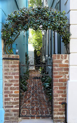 Visible from the street, a weathered wrought iron gate between buildings guards a walkway lined with plants and old brick pavers. The gate is crowned with an arch of evergreen star jasmine.