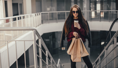 Young pretty woman blogger with smartphone, cup of coffee and shopping bags in city mall