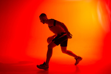 Fototapeta na wymiar Young athletic man, basketball player dribbling with ball on red and orange background