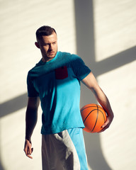 Young athletic man, basketball player holding ball and standing near wall with shadows from window