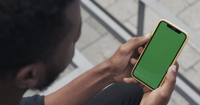 Over sholder view of african male holding smartphone vertically with mock up green screen. View from above of man sitting and looking at phone screen outdoors. Chroma key. Concept of tech.