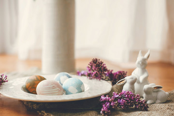 Obraz na płótnie Canvas Stylish Easter eggs on vintage plate, white bunny rabbits and lilac flowers on rustic fabric on wooden table. Rural easter composition of natural dyed eggs and spring purple flowers