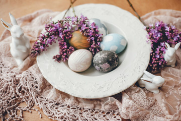 Stylish Easter eggs on vintage plate, white bunnies and lilac flowers on fabric on wooden table. Rural composition of colorful natural dyed easter eggs and spring flowers. Holiday decor