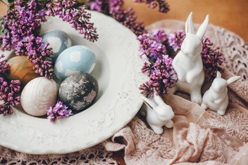 Obraz na płótnie Canvas Stylish Easter white bunnies, modern easter eggs on vintage plate, and lilac flowers on rural fabric. Natural dyed easter eggs and spring flowers. Space for text. Holiday decor