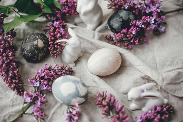 Obraz na płótnie Canvas Stylish Easter eggs, white bunnies and lilac flowers on linen rural fabric. Modern holiday table decor. Happy Easter. Natural dyed easter eggs and spring flowers