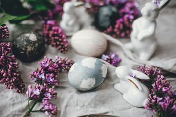 Obraz na płótnie Canvas Happy Easter. Rustic composition of natural dyed easter eggs, lilac flowers and bunny rabbits on linen fabric on wooden table. White ceramic bunny and spring flowers.