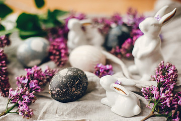 Happy Easter. Modern Easter eggs, white bunnies and lilac flowers on linen rural fabric. Stylish holiday table decor. Space for text. Natural dyed easter eggs and spring flowers.
