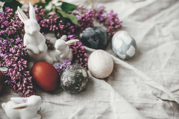 Modern Easter eggs, white bunnies and lilac flowers on linen rural fabric. Stylish holiday table decor. Happy Easter. Space for text. Natural dyed easter eggs and spring flowers.