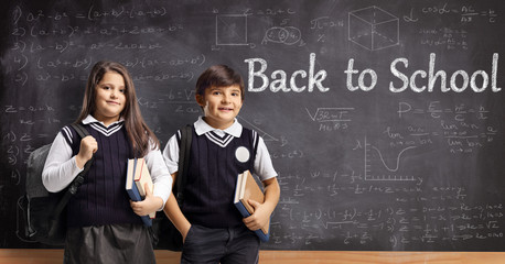 Boy and girl in uniforms standing in front of a chalkboard with math formulas and written text back to school