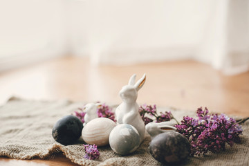 Obraz na płótnie Canvas Modern Easter eggs, white bunnies and lilac flowers on rustic fabric. Stylish holiday table decor. Space for text. Happy Easter. Natural dyed easter eggs and spring flowers. Rural still life