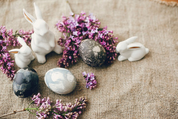 Modern Easter eggs, white bunnies and lilac flowers on rustic fabric. Stylish holiday table decor. Space for text. Happy Easter. Natural dyed easter eggs and spring flowers. Rural still life
