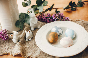 Stylish Easter eggs on vintage plate, white bunny rabbits and lilac flowers on rustic fabric on wooden table. Rural easter composition of natural dyed eggs and spring purple flowers