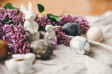 Modern Easter eggs, white bunnies and lilac flowers on linen rural fabric. Stylish holiday table decor. Happy Easter. Space for text. Natural dyed easter eggs and spring flowers.