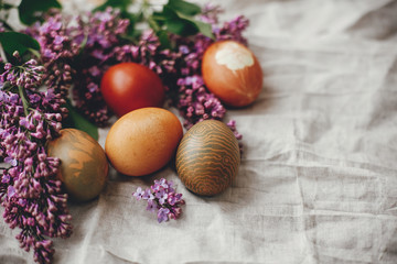 Fototapeta na wymiar Modern colorful Easter eggs on rustic linen fabric and purple lilac flowers. Rural still life. Happy Easter. Space for text. Natural dyed easter eggs. Stylish holiday table decor