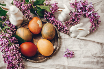Obraz na płótnie Canvas Stylish Easter eggs on rustic plate, white bunnies and lilac flowers on linen rural fabric. Top view. Natural dyed yellow easter eggs and spring flowers. Holiday decor. Happy Easter