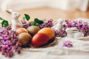 Obraz na płótnie Canvas Happy Easter. Stylish Easter eggs on rustic plate, white bunnies and lilac flowers on linen rural fabric. Natural dyed yellow easter eggs and spring flowers. Holiday decor.