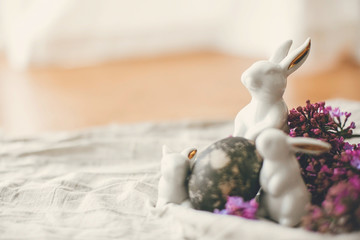 Happy Easter. Modern white bunnies, Easter egg and lilac flowers on rustic fabric. Space for text. Rural still life. Natural dyed easter eggs and spring flowers. Stylish holiday table decor