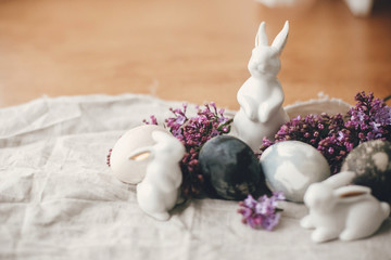 Obraz na płótnie Canvas Modern Easter eggs, white bunnies and lilac flowers on linen rustic fabric. Stylish holiday table decor. Space for text. Happy Easter. Natural dyed easter eggs and spring flowers.