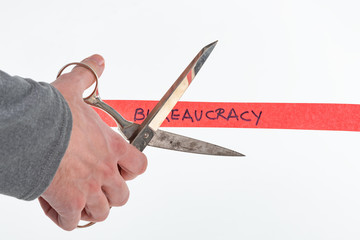 Male hand cutting through bureaucracy red tape with scissors isolated on white with a shallow depth of field and copy space - obrazy, fototapety, plakaty
