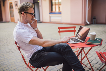 Pleased man speaking on smartphone and using laptop in street cafe