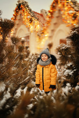 cute boy standing between pine trees on the street decorated with christmas lights at winter time