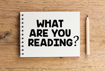 The question What Are You Reading written in an office notebook set on a wooden table. Top view, high resolution photography.