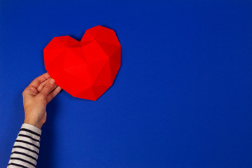 Female hand holding red polygonal heart on blue background. Top view