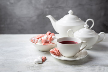 Beautiful romantic breakfast with red tea Hibiscus and meringue cookies in the shape of hearts and candy. White tea-set stand on a wooden table with a concrete background. Still life with copy space.