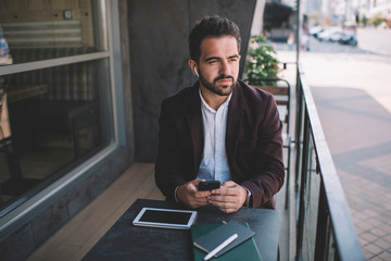  Caucasian male millennial sitting on terrace in cafe using earbuds for listening audio book about business development, confident formally dressed male using modern devices for listening music .