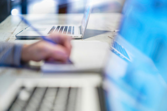 Closeup of hand woman writing working with a pen on a document, close to her computer laptop pc in her office on her workplace, desktop career screen notebook business, cold blue atmosphere blurred