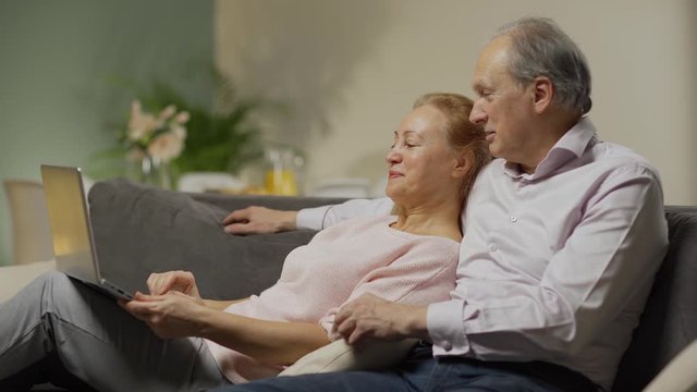 Side view of senior couple relaxing on sofa at home, looking and pointing at laptop screen, discussing and choosing items in online shop or browsing through pictures on Internet