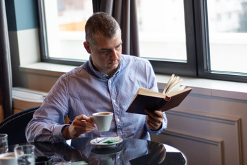 Elegant man reading a book and drinking coffee on the table in a restaurant. Man wears a modern shirt, relaxing and enjoying. Education concept. Close up, selective focus
