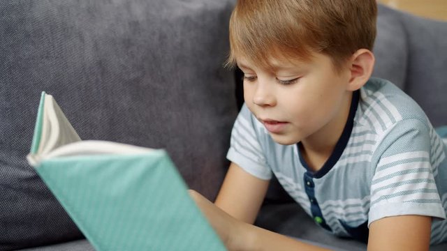 Handsome young boy is reading book at home learning relaxing on couch in apartment enjoying self education. Smart children, literature and literacy concept.