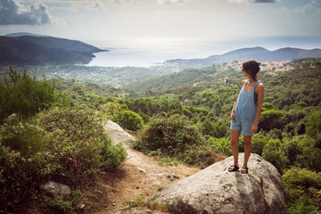 Young woman on holiday wearing denim summer dungaree shorts and sandals admiring panorama from hill...