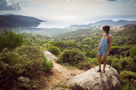 Young woman on holiday wearing denim summer dungaree shorts and sandals admiring panorama from hill overseeing bay in Tuscany, Italy