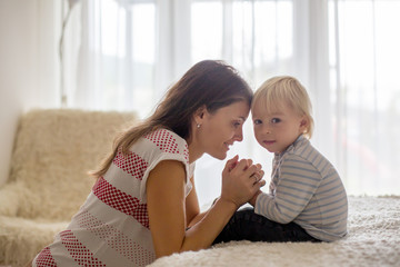 Mother and toddler son, praying at home, sitting on bed in bedroom