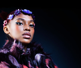 young pretty african american woman in spotted fur coat and flowers on head smiling on black background, fashion people concept