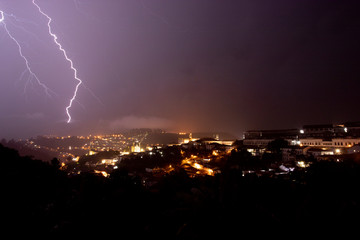 Naklejka na ściany i meble Lightning ray finding its way through the pouring rain striking in the hills behind the historic urban centre of Ouro Preto in Brazil lighting up the night sky with city lights in the foreground