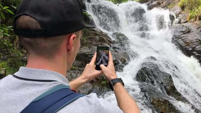 Travel man in shirt and cap on his head, with backpack on the back, take photo of Datanla Waterfall on his phone, Vietnam, Southeast Asia.