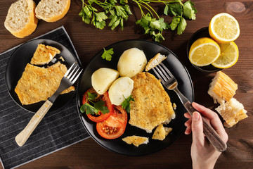Hand of a person who is eating a delicious baked battered fish seasoning with fresh parsley and lemon and accompanied by cooked potatoes, fresh tomato and freshly baked bread. On a rustic wooden table