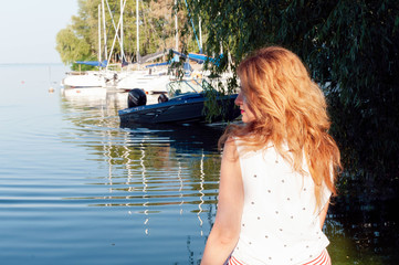 Fototapeta premium back view of beautiful woman with long hair in white blouse sitting near river water with boats and looking away in yachts club