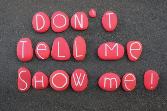 Don't tell me, show me. Motivational phrase composed with red colored and carved stone letters over black volcanic sand