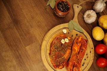 raw meat, two meat pickled in spices meat medallions on a round wooden board on the table, garlic, bay leaf, potatoes and tomatoes nearby, cooking concept, copy space