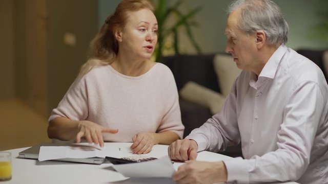 Unhappy elderly couple getting stressed while calculating finances together sitting at table at home and looking at paper bills with expenses