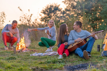 Group of happy friends with guitar, having fun outdoor, near bonfire and tourist tent. Camping fun happy family