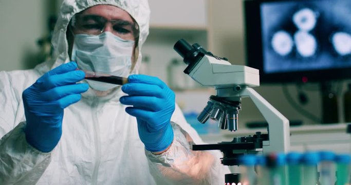 A laboratory technician working in a lab examines a blood sample as an image of the Wuhan coronavirus virus is displayed on a monitor in the background.