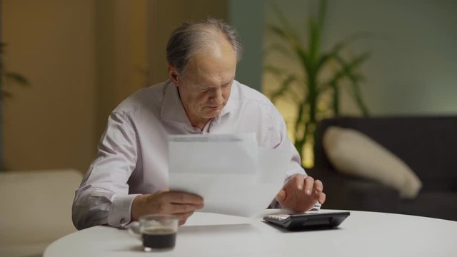 Senior man getting stressed and putting head in hands after looking at paper bills and calculating expenses sitting at table at home